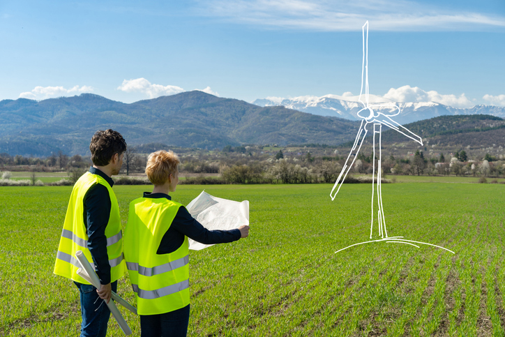 Concept of a large wind turbine drawn onto a field, with two people wearing reflective jackets looking at blueprints, under a blue sky with mountains in the distance
