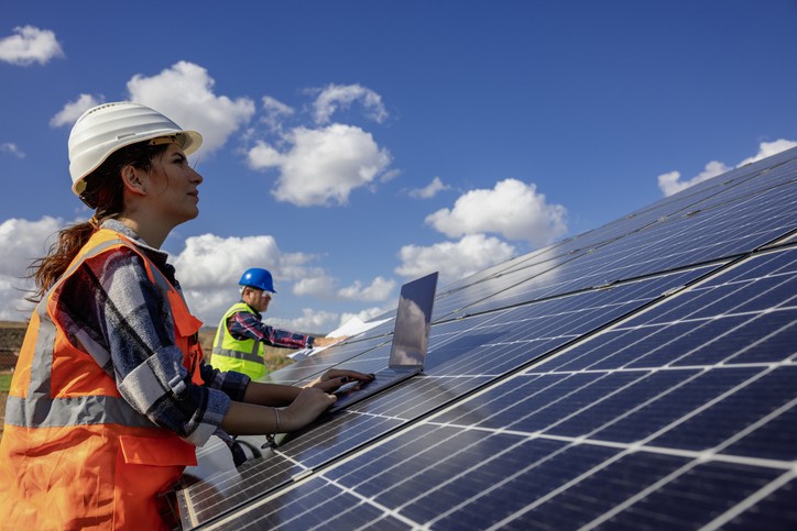 Female tradesperson leaning on a solar panel with blue sky in the background.
