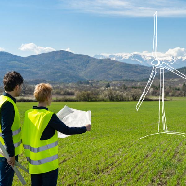 Concept of a large wind turbine drawn onto a field, with two people wearing reflective jackets looking at blueprints, under a blue sky with mountains in the distance